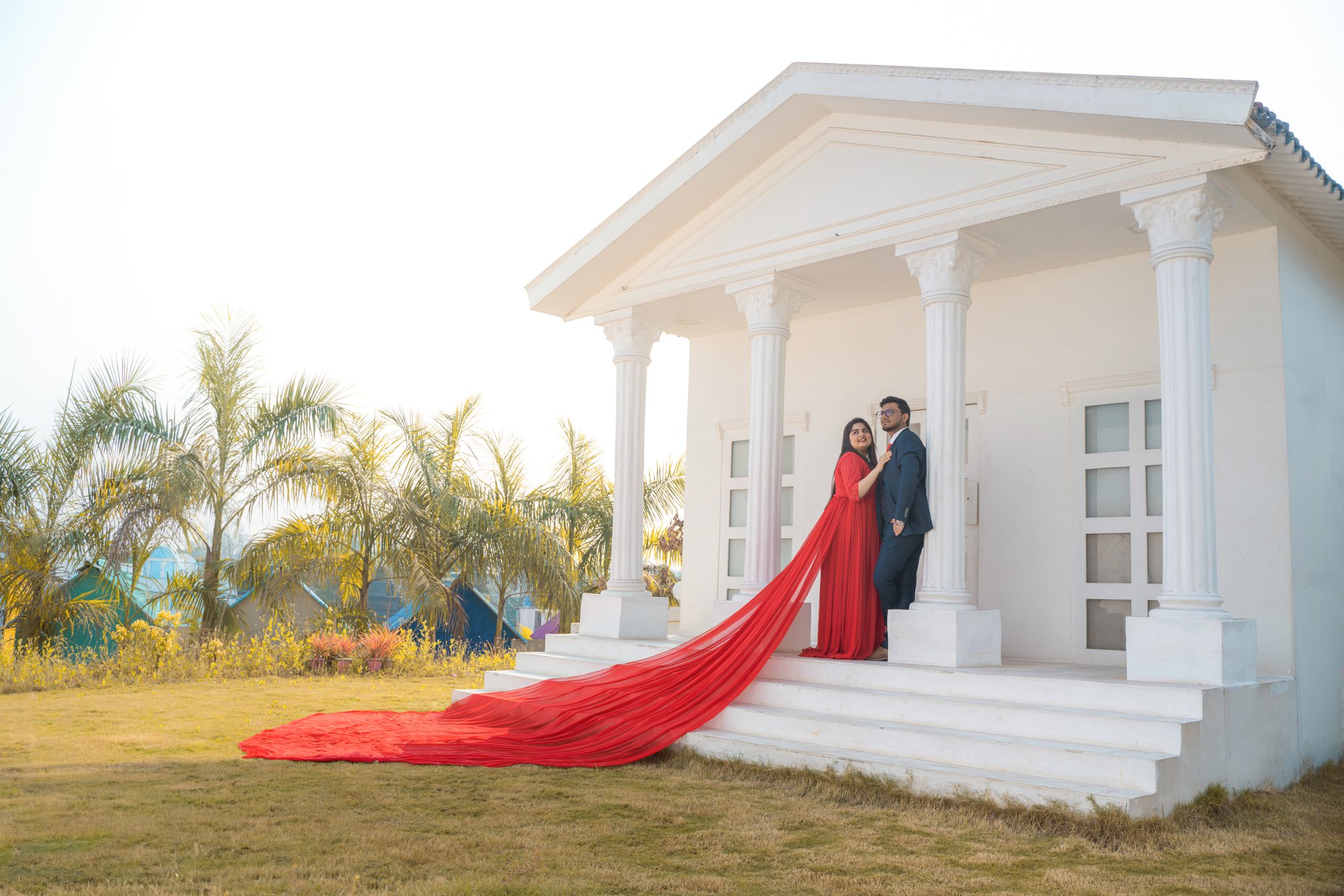 A dreamy pre-wedding moment—groom in a sharp suit and bride in red standing in a white house.